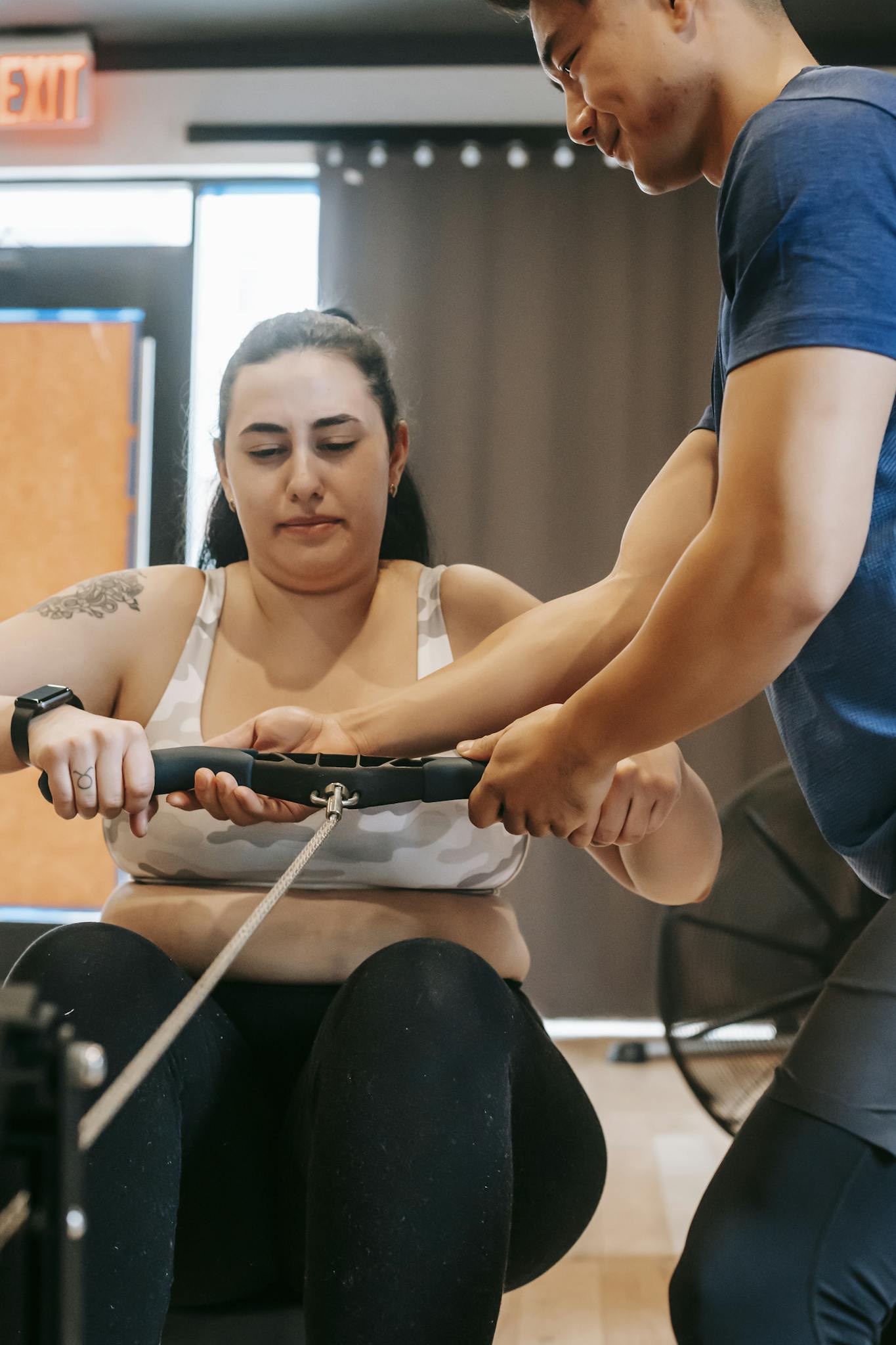 A personal trainer supports a woman during an intense rowing workout session indoors.